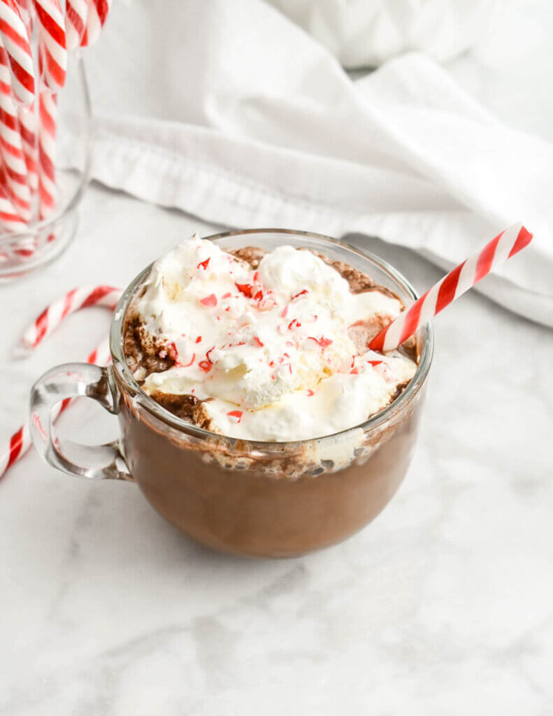 A peppermint mocha in a clear mug topped with whipped cream, crushed candy cane and a candy cane stick set on a marble countertop with a white napkin and candycanes around it.