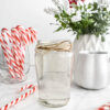 A glass jar with peppermint simple syrup on a grey marble countertop with candycanes, a cup of coffee, and a white vase with red and white flowers in the background.