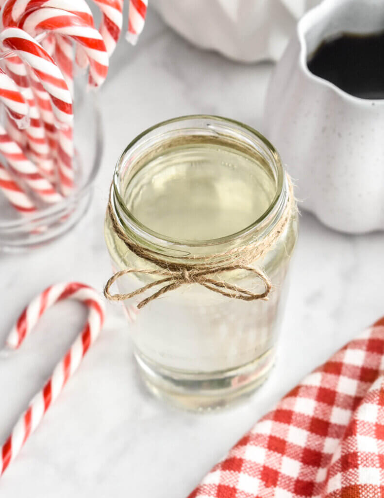 A glass jar of peppermint syrup on a grey marble countertop surrounded by candycanes, a cup of coffee and a red and white checkered napkin.