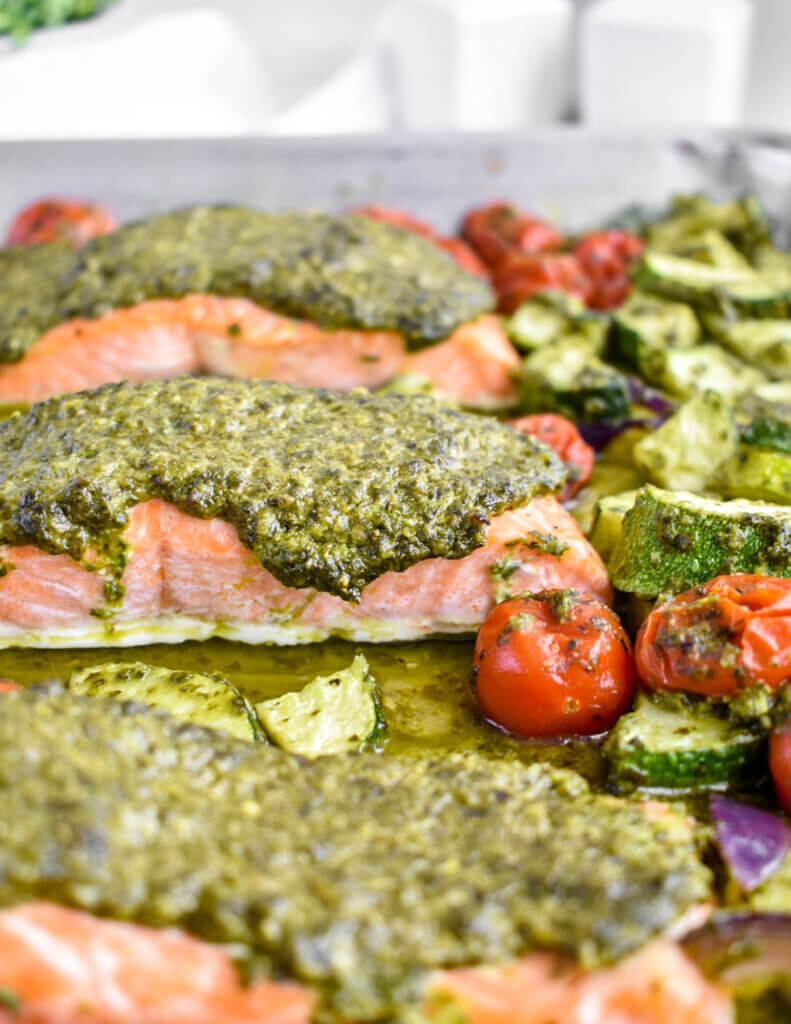 Close up of baked salmon topped with pesto and baked with cherry tomatoes and zucchini in a sheet pan.
