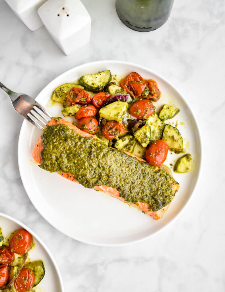 Pesto topped salmon and pesto coated zucchini and cherry tomatoes on a white dinner plate set on a grey marble countertop.