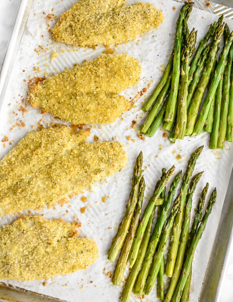 Fillets of crispy breaded fish and asparagus on a parchment lined sheet pan.