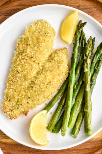 Crispy oven baked breaded fish and asparagus served with lemon wedges on a white plate.