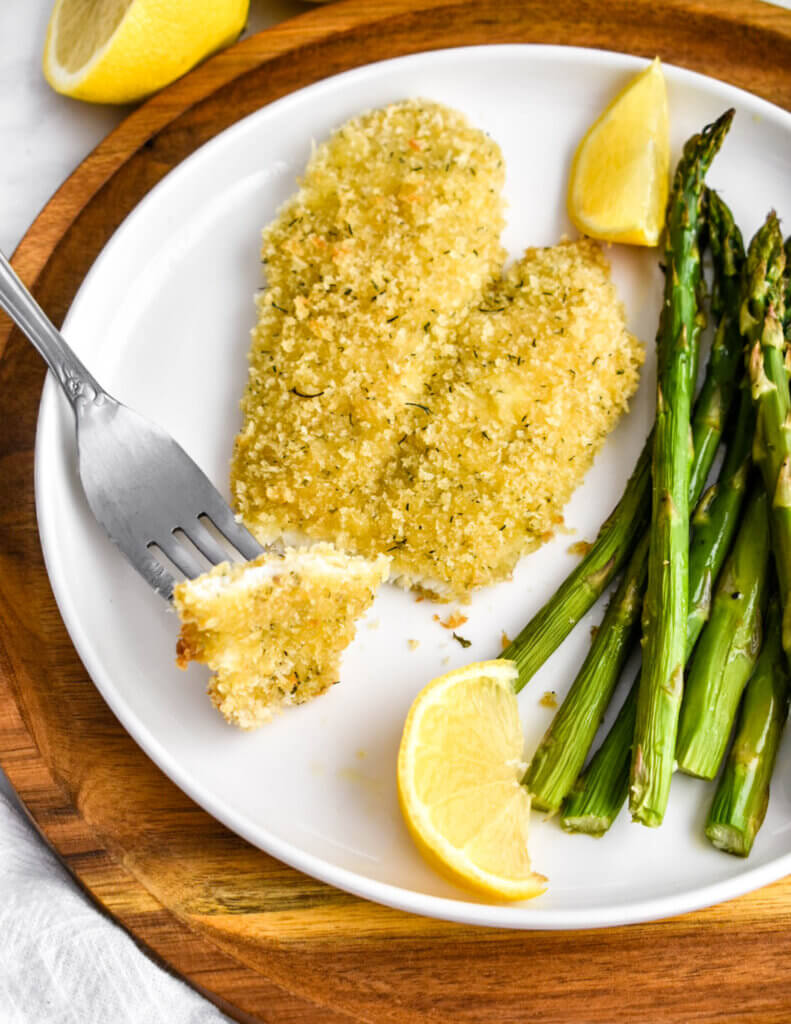 Closeup of a breaded fish fillet with a bite speared onto a fork on a plate served with asparagus and lemon wedges.