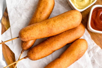 4 corn dogs on a wood platter lined with parchment paper served with two small bowls containing mustard and ketchup.