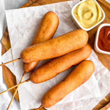 4 corn dogs on a wood platter lined with parchment paper served with two small bowls containing mustard and ketchup.