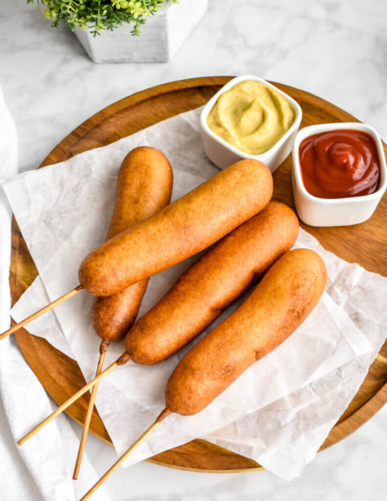Corn dogs on a parchment lined wooden platter served with two dipping bowls filled with mustard and ketchup.