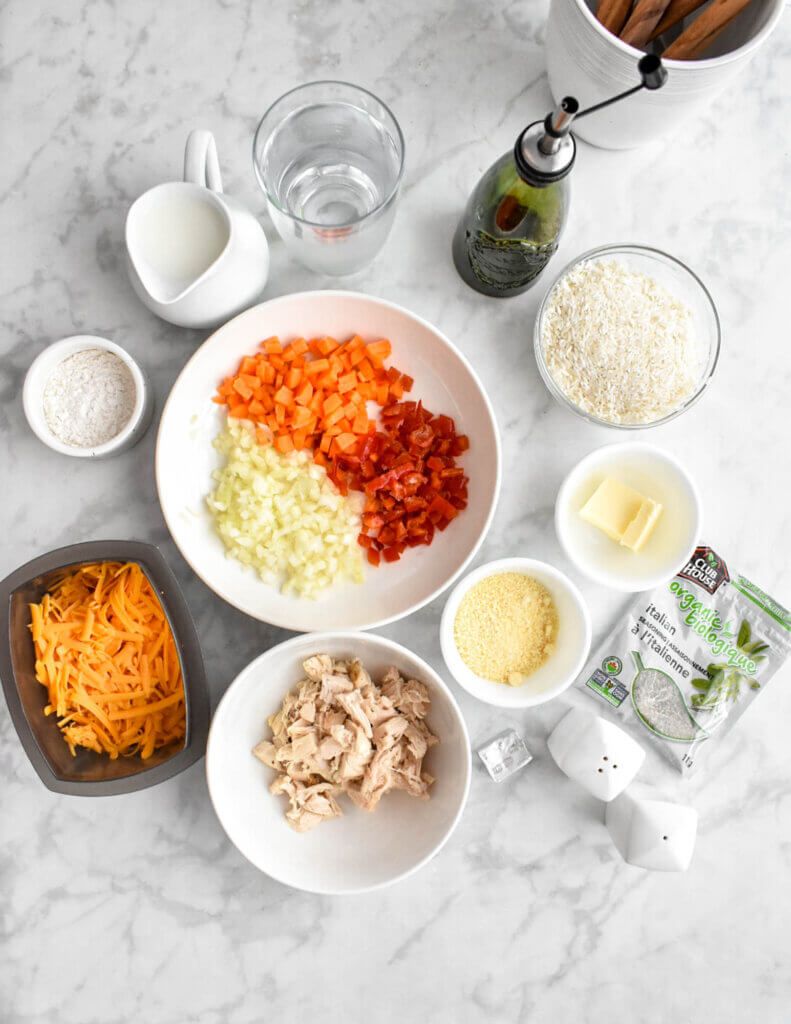 Ingredients for the casserole including rice, chicken, chopped carrots, onions and peppers, cheese, milk, oil and seasoning set on a grey marble countertop.