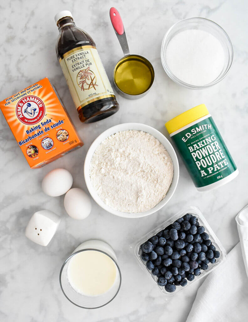 The ingredients for Buttermilk Blueberry Muffins including flour, buttermilk, blueberries, melted butter, eggs, vanilla, salt, sugar, baking soda, and baking powder on a grey marble counter top.