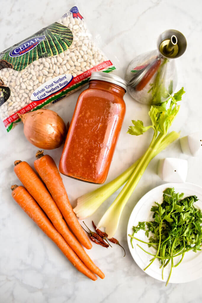 Ingredients in Fasolada including dried white beans, onion, 3 carrots, 2 celery ribs, parsley, olive oil, tomato sauce, and dry red chilis on a grey marble counter top.