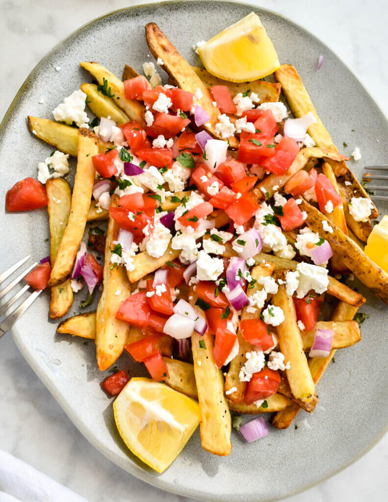 Closeup of a platter of french fries topped with diced tomato, onion, crumbled feta and served with lemon wedges.