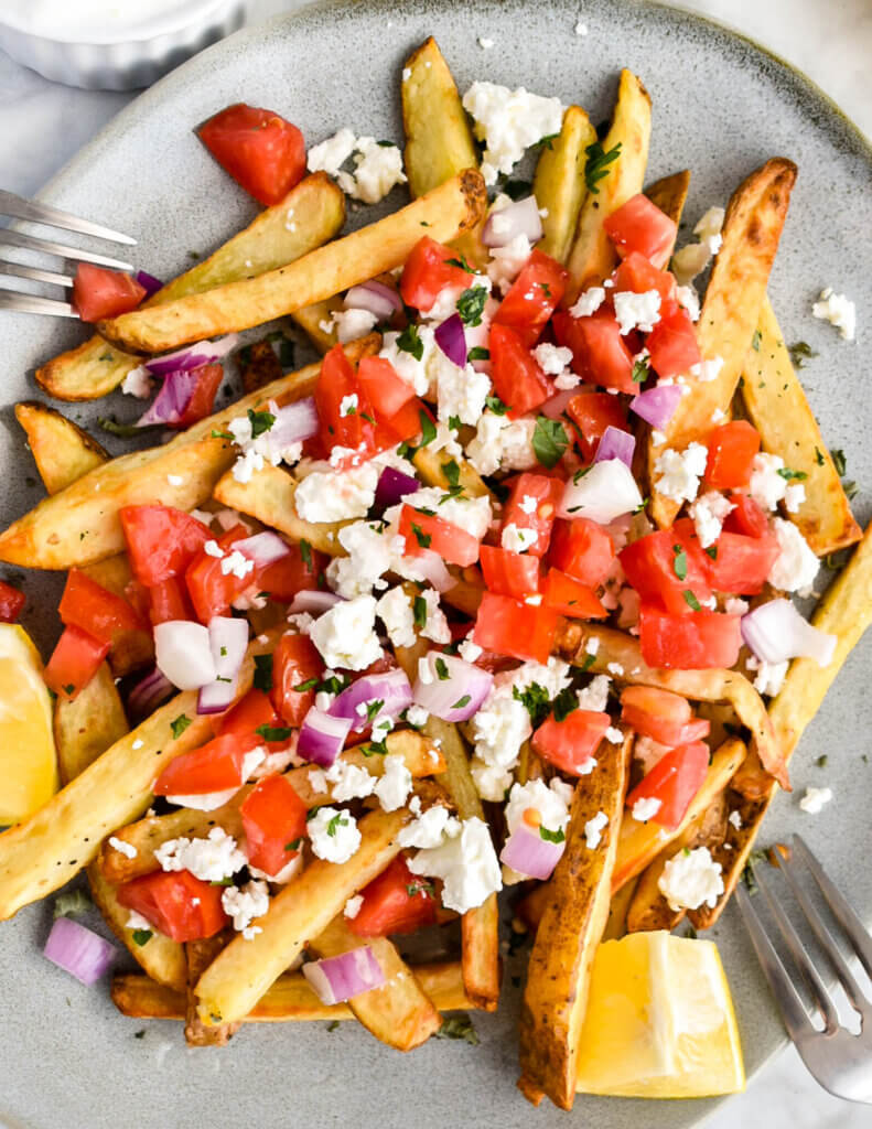 Closeup of a platter of Loaded Greek Feta Fries topped with feta, onion, tomato and served with lemon wedges.