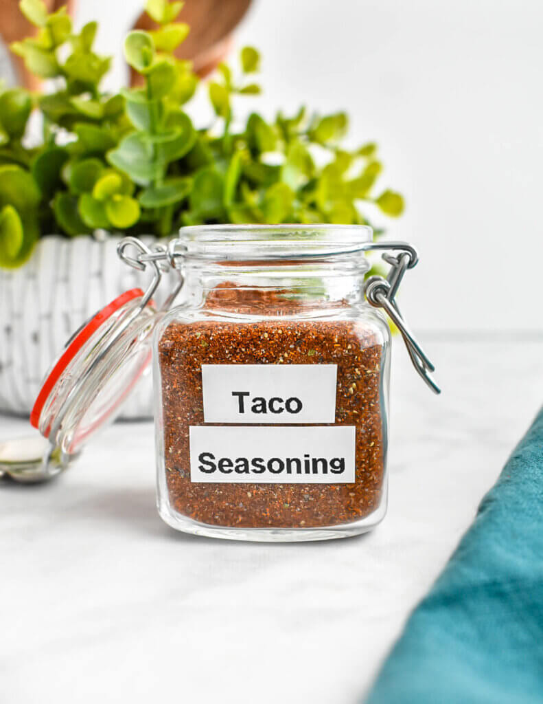 A jar of taco seasoning with a printed "Taco Seasoning" label set on a grey marble counter next to a green plant.