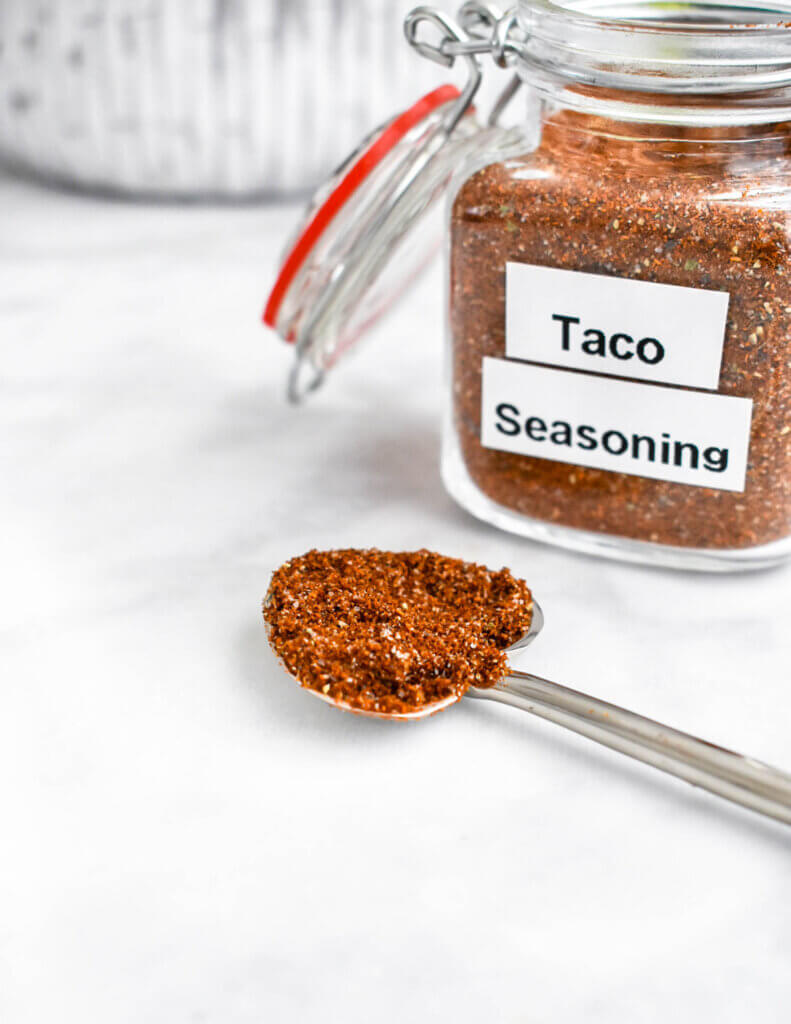 A spoonful of taco seasoning set on a grey marble counter in front of a jar of taco seasoning.