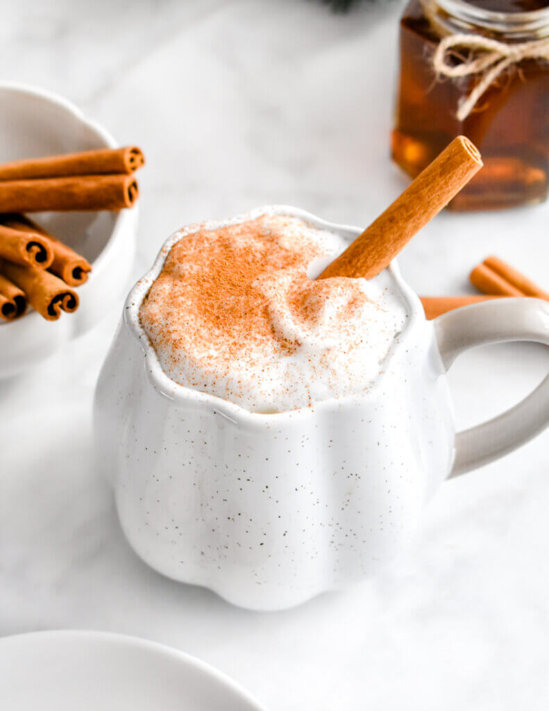 Closeup of a mug of cinnamon dolce latte set next to a bowl of cinnamon sticks and a jar of cinnamon syrup.