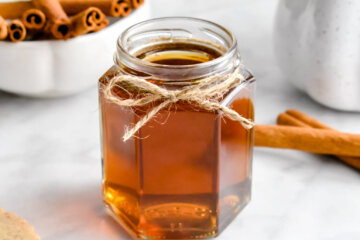 A jar of Cinnamon Simple Syrup set on a grey marble counter next to cinnamon sticks and a mug of coffee.