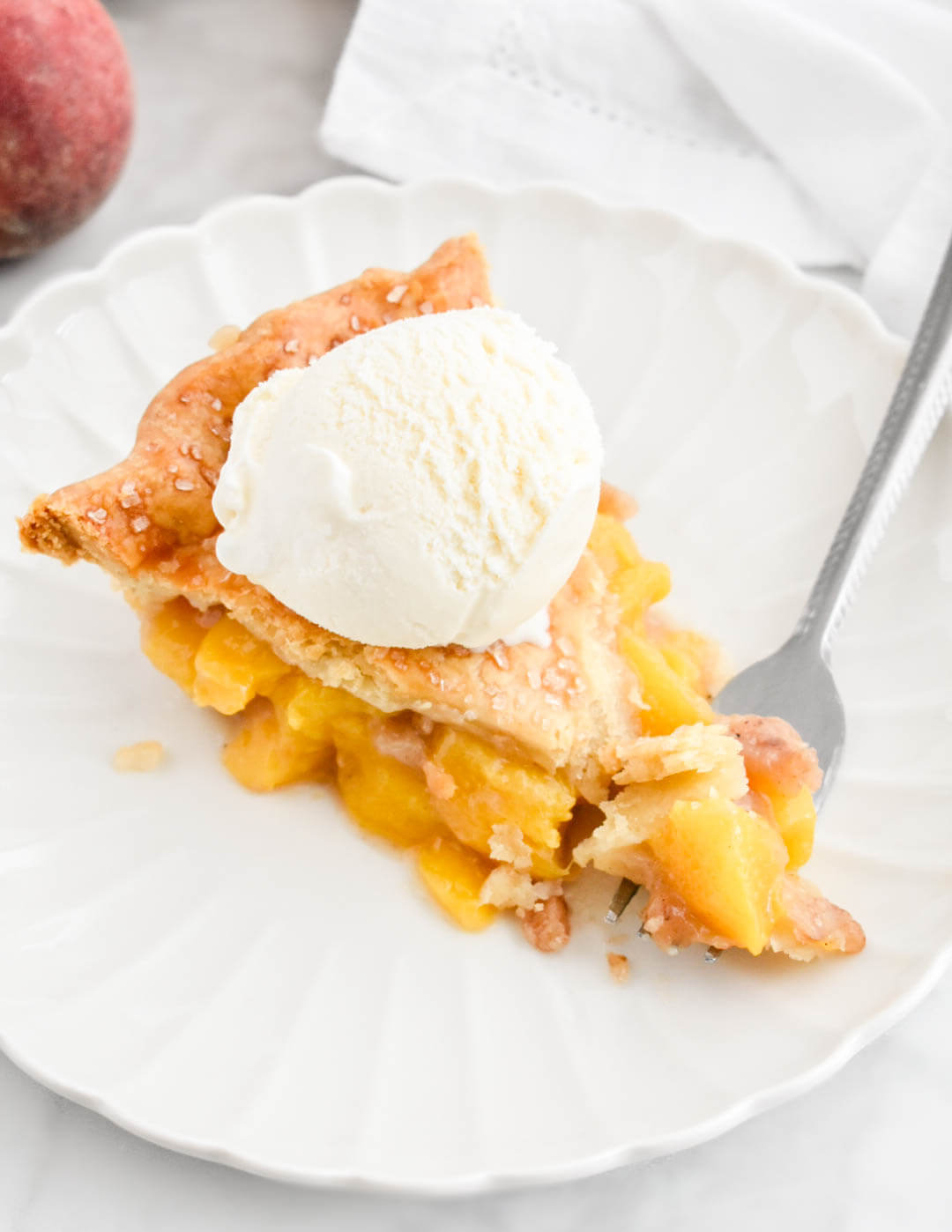 A fork being cut into a slice of peach pie topped with vanilla ice cream.