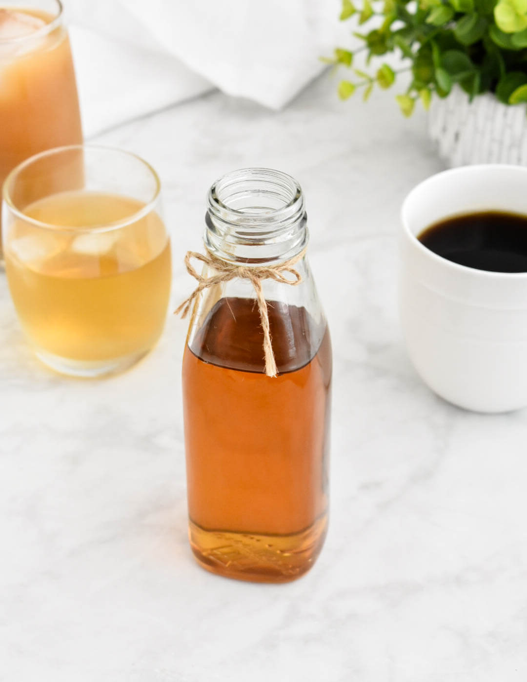 A bottle of vanilla simple syrup set on a grey counter next to a cocktail, cup of coffee, and iced coffee.