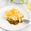 A slice of beef pie with filo topping on a white plate set next to a glass of white wine.