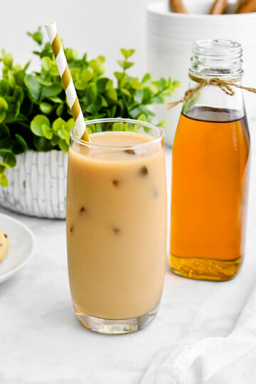 An iced vanilla latte set on a grey counter next to a green plant and a bottle of vanilla syrup.