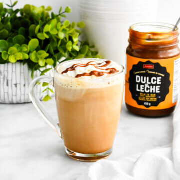 Dulce de leche latte in a clear mug set down next to a jar of dulce de leche.