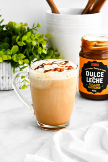 Dulce de leche latte in a clear mug set down next to a jar of dulce de leche.