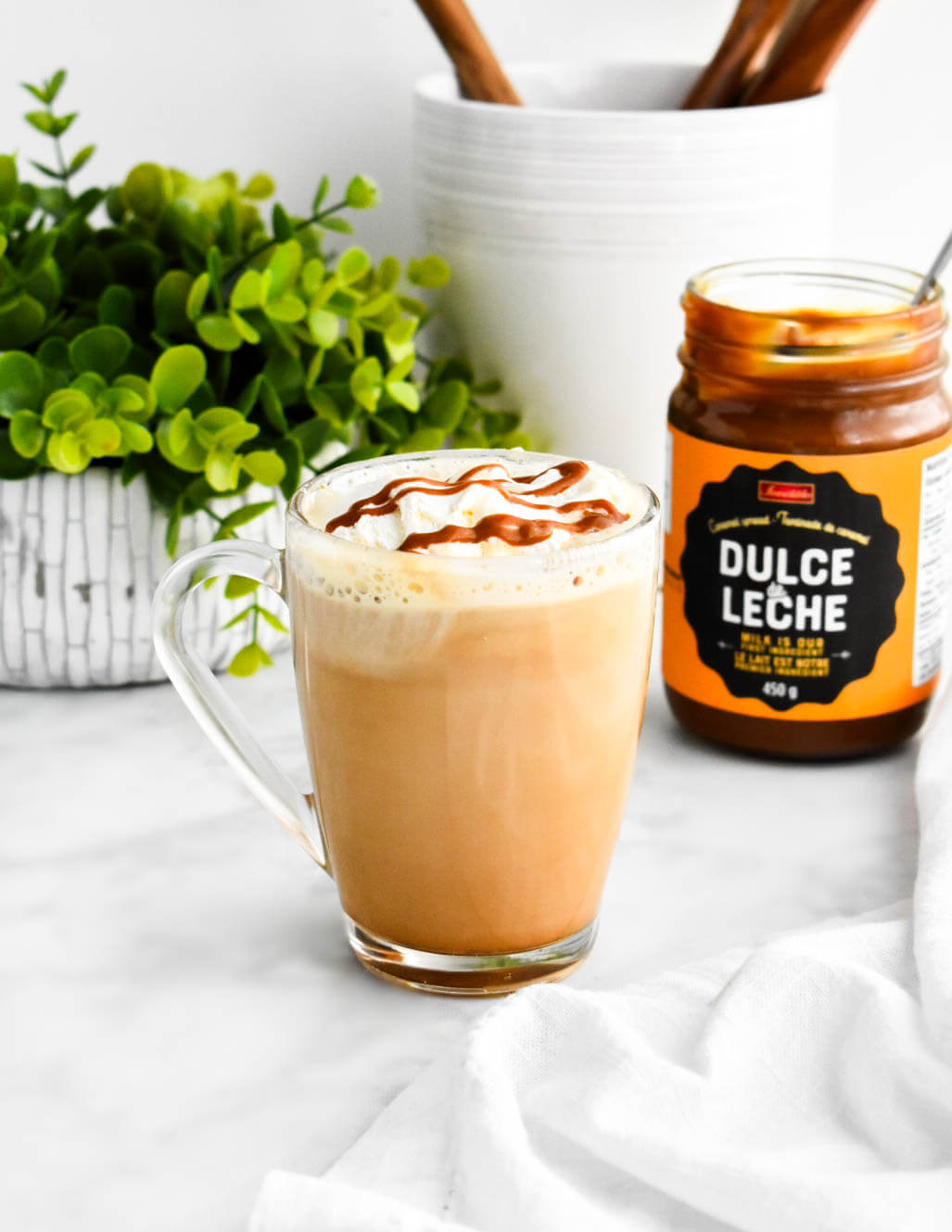 Dulce de leche latte in a clear mug set down next to a jar of dulce de leche.