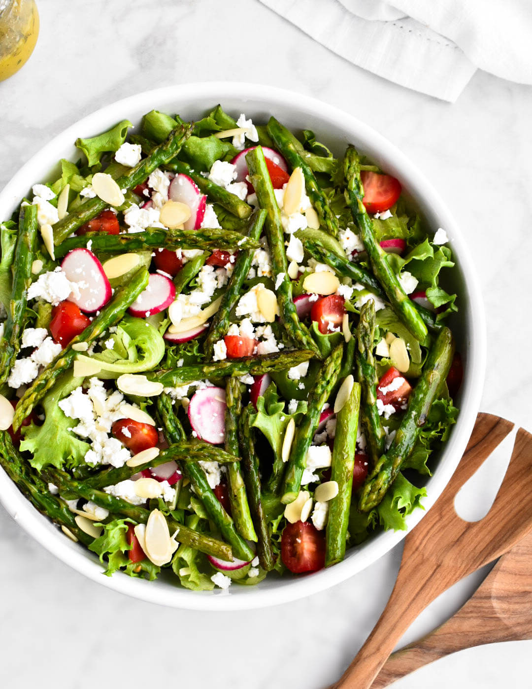 Asparagus salad in a white serving bowl set next to a jar of lemon herb vinaigrette and wooden salad servers.