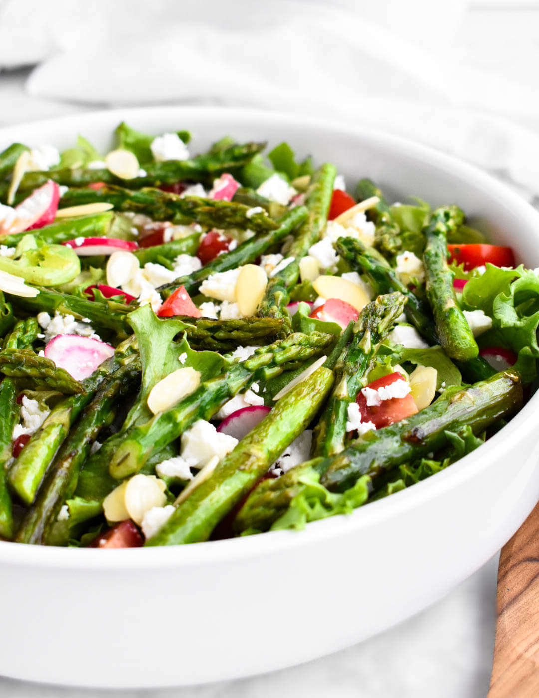 Closeup of a salad made of greens, asparagus, radishes, cherry tomatoes, feta, and almond slices.