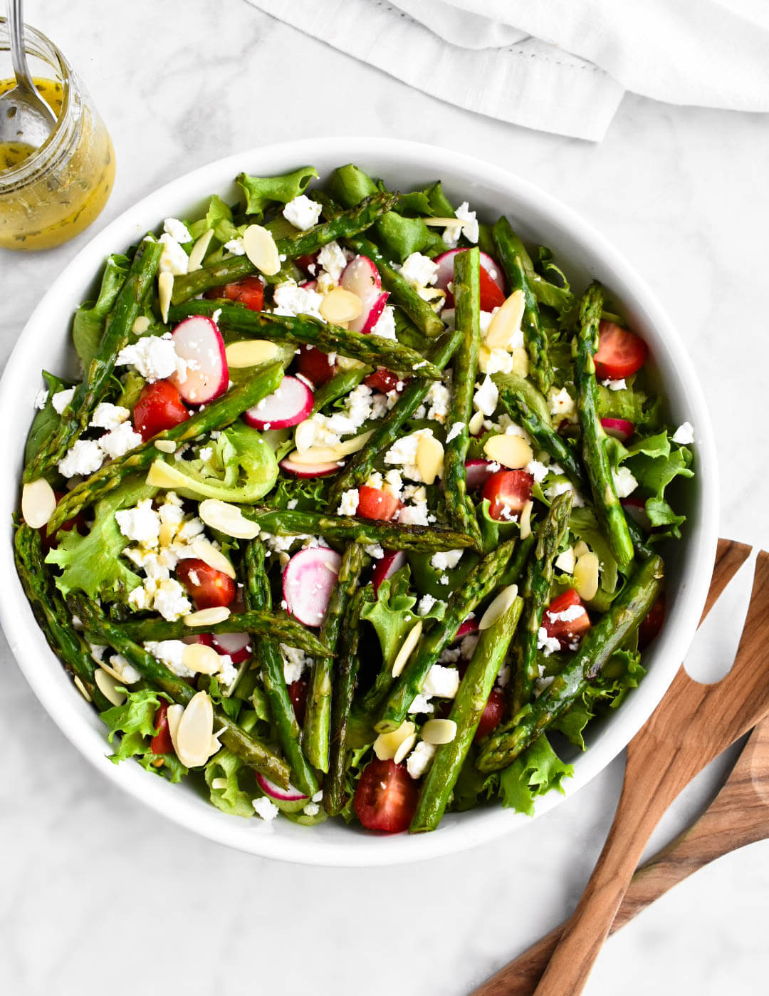 Asparagus salad in a white serving bowl set next to a jar of lemon herb vinaigrette and wooden salad servers.