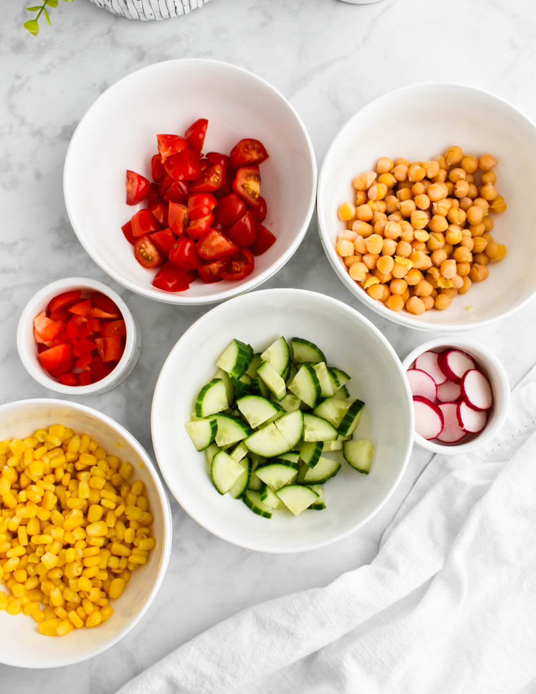 Chopped tomatoes, cucumbers, radishes, and red pepper in bowls along with bowls of corn kernels and chickpeas.